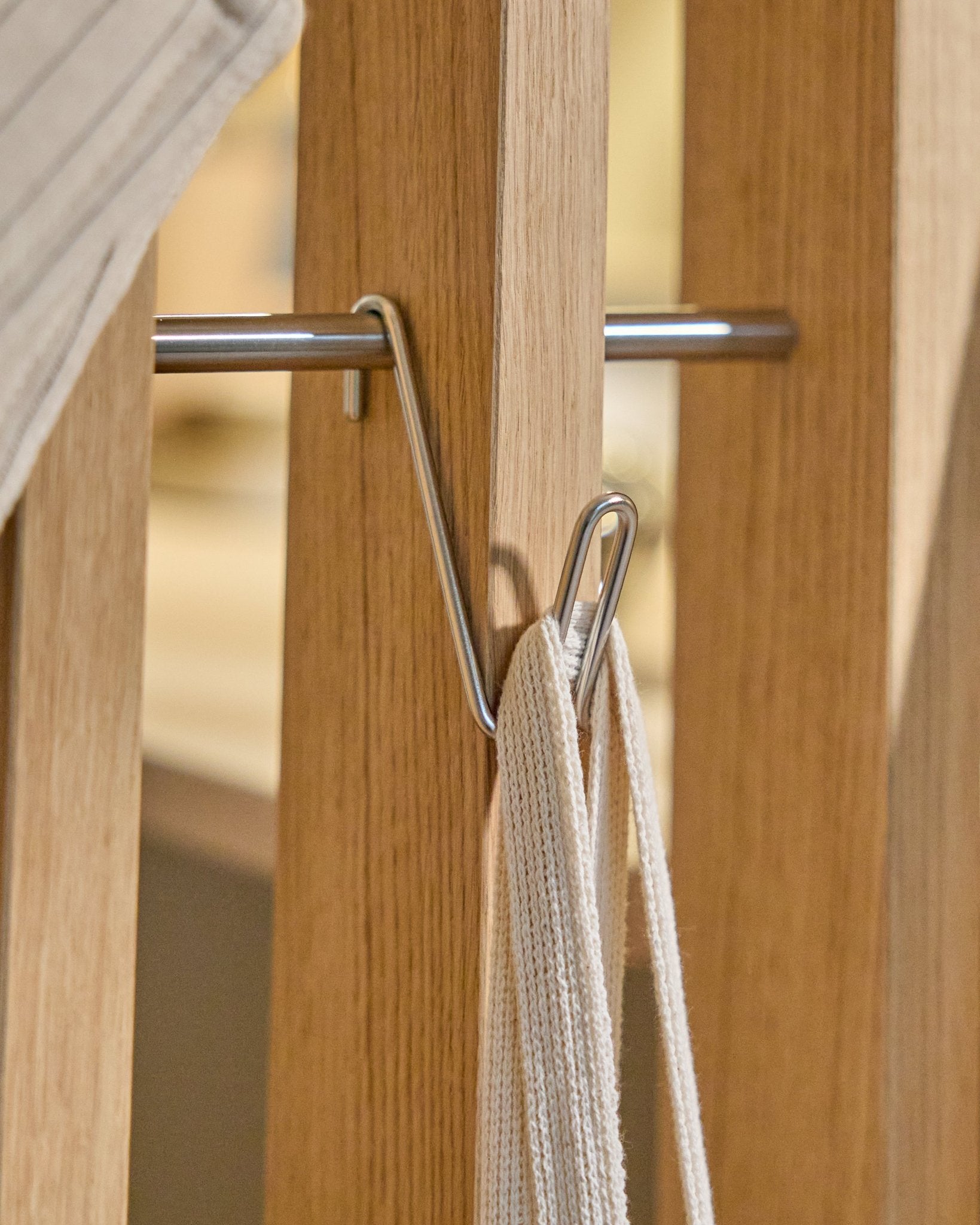 Le Alva Wire Hook / BM1 (acier inoxydable Steel, format court) de WoodUpp est monté sur un rail en bois Panneau acoustique et supporte une sangle blanche avec tige métallique ; fond beige.